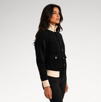 A woman modeling a black jacket with contrasting cream ribbed trim at the collar, cuffs, and hem, paired with black pants against a light gray studio background.