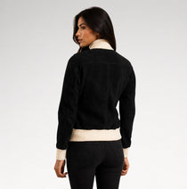 A woman shown from the back wearing a black suede-like jacket with contrasting cream ribbed trim at the collar, cuffs, and hem, paired with black jeans against a light gray studio background.