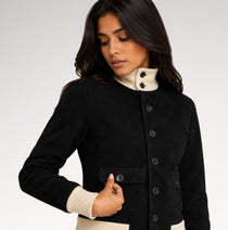 A woman wearing a black jacket with a cream ribbed mock neck and cuffs, shown against a light gray studio background.