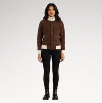 A woman modeling a brown suede jacket with cream ribbed trim and a high cream collar, paired with black pants and black boots, against a light gray studio background.