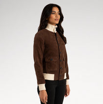 A woman modeling a brown suede button-up cardigan with cream ribbed trim, worn over a cream mock-neck top and black pants, against a light gray studio background.