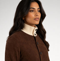 A woman modeling a brown suede jacket with cream ribbed trim at the mock neck, cuffs, and hem, paired with black pants against a light gray studio background.