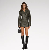 A model stands against a light gray studio background wearing an olive green faux-leather double-breasted jacket and matching mini skirt, paired with black knee-high boots and holding a small black handbag.