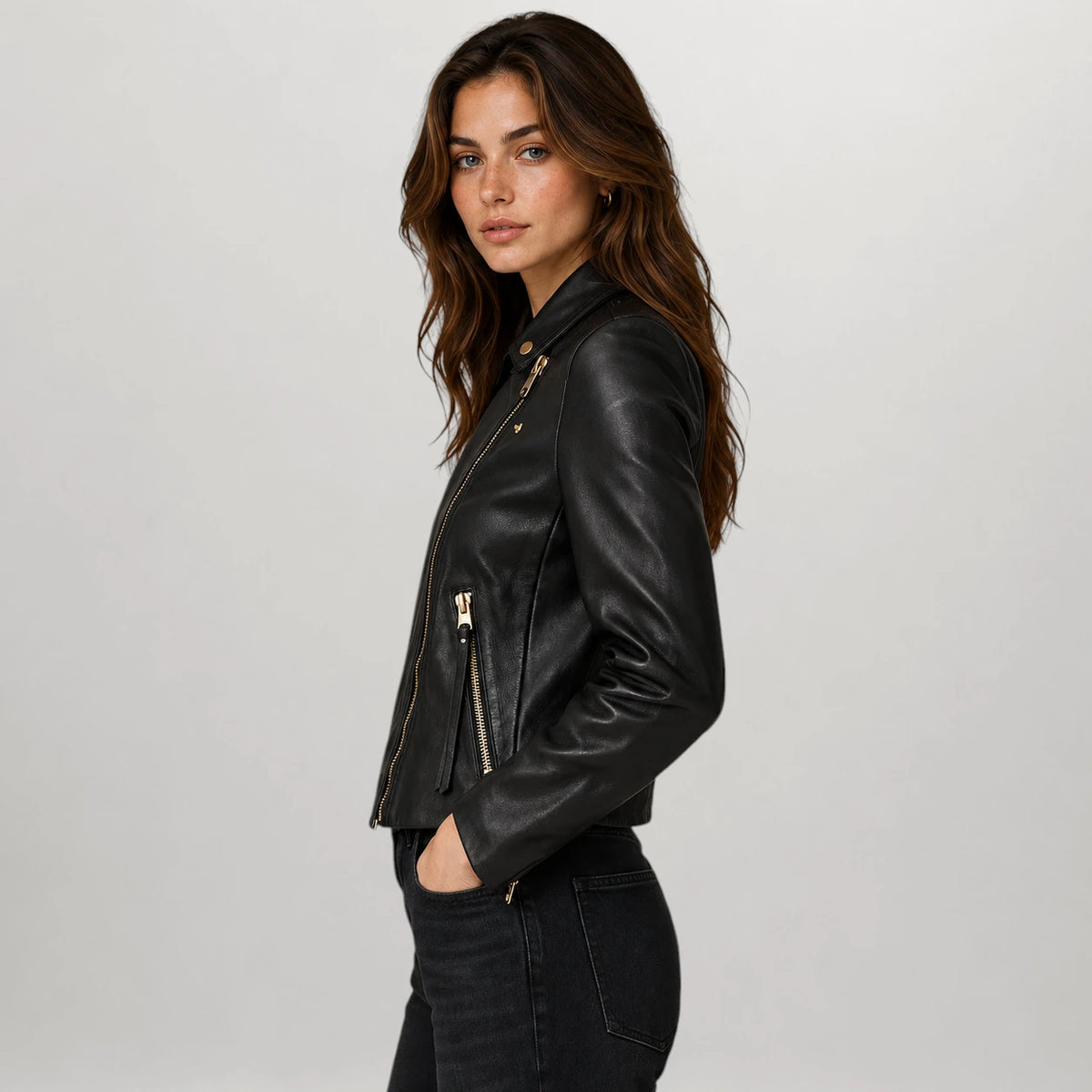 A woman wearing a black leather moto jacket with gold-tone zippers, shown in a side profile against a light gray studio background.