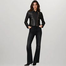 A woman modeling a black leather moto jacket with gold-tone zippers, paired with black jeans and black heels, against a light gray studio background.