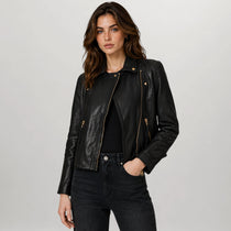 A woman modeling a black leather moto jacket with gold-tone hardware, paired with a black top and dark jeans, against a light gray studio background.