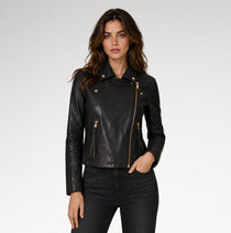 A woman modeling a black faux-leather moto jacket with gold-tone hardware, paired with dark gray jeans, against a light gray studio background.