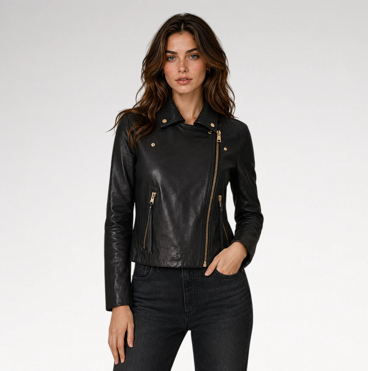 A woman modeling a black faux-leather moto jacket with gold-tone hardware, paired with dark gray jeans, against a light gray studio background.