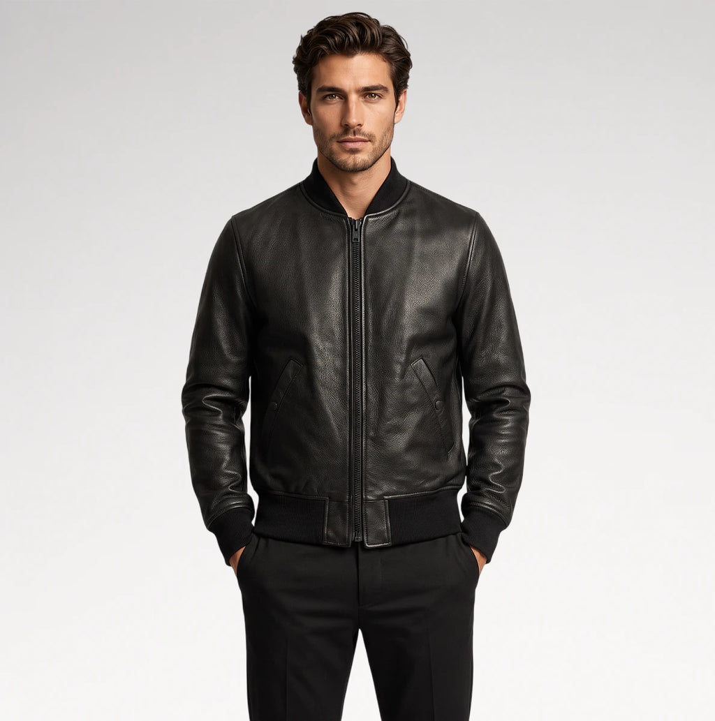 A man wearing a black leather bomber jacket with a front zipper, ribbed collar, cuffs, and hem, paired with black trousers, photographed against a light gray studio background.