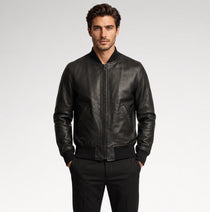 A man wearing a black leather bomber jacket with a front zipper, ribbed collar, cuffs, and hem, paired with black trousers, photographed against a light gray studio background.