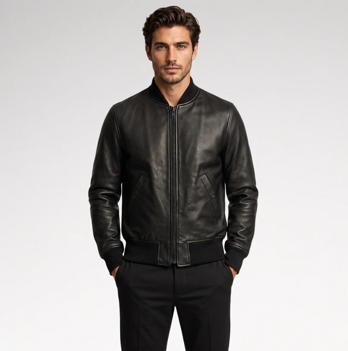 A man wearing a black leather bomber jacket with a front zipper, ribbed collar, cuffs, and hem, paired with black trousers, photographed against a light gray studio background.