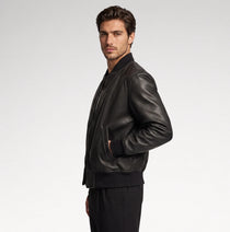A man wearing a black leather bomber jacket and black pants, shown in profile against a light gray studio background.
