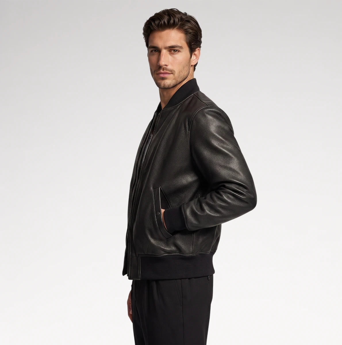 A man wearing a black leather bomber jacket and black pants, shown in profile against a light gray studio background.