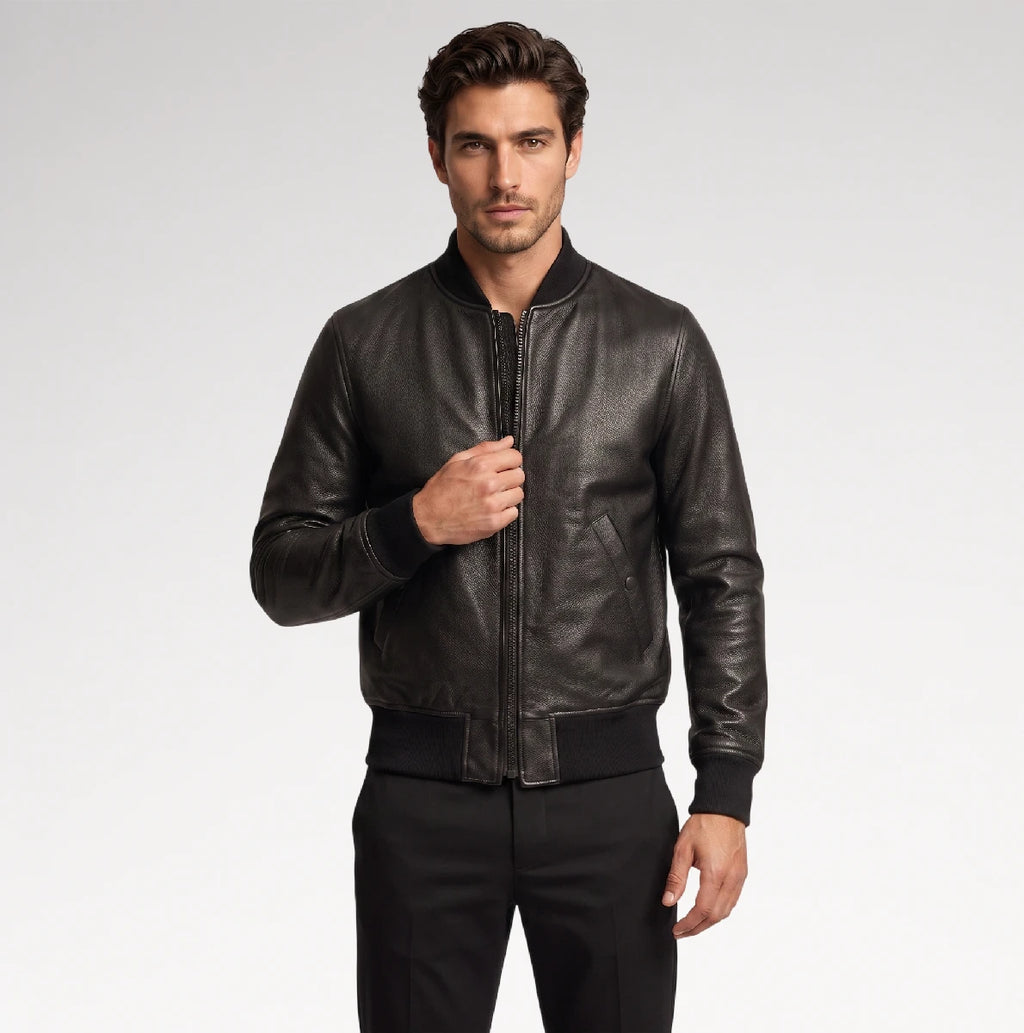 A man wearing a black leather bomber jacket with a front zipper, ribbed collar, cuffs, and hem, paired with black trousers, photographed against a light gray studio background.