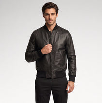 A man wearing a black leather bomber jacket with a front zipper, ribbed collar, cuffs, and hem, paired with black trousers, photographed against a light gray studio background.
