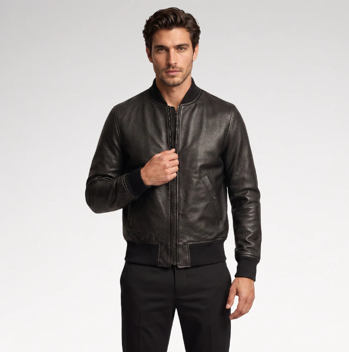 A man wearing a black leather bomber jacket with a front zipper, ribbed collar, cuffs, and hem, paired with black trousers, photographed against a light gray studio background.