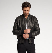 A man wearing a black leather bomber jacket over a white t-shirt and black pants, standing against a light gray studio background.