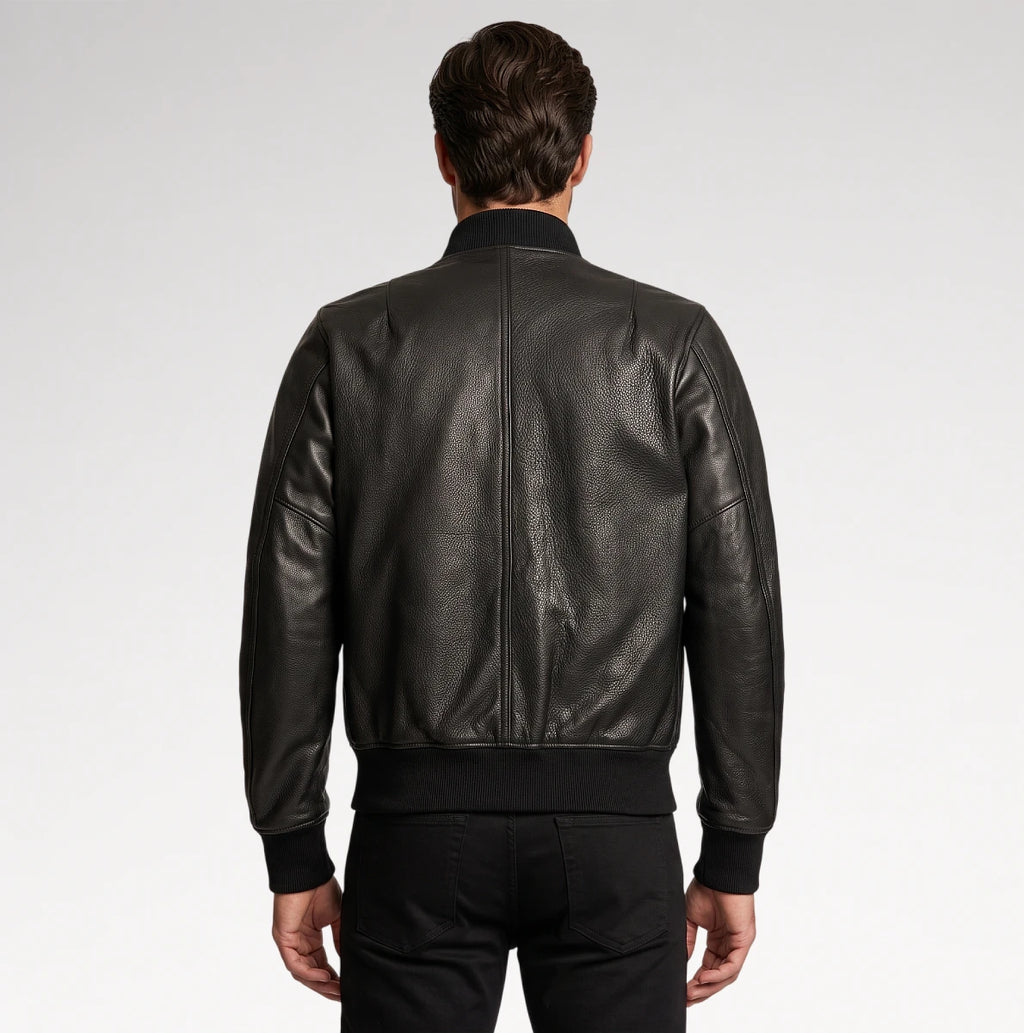 A man viewed from the back wearing a black leather bomber jacket with a ribbed collar, cuffs, and hem, paired with black pants, against a light gray studio background.
