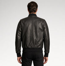 A man viewed from the back wearing a black leather bomber jacket with a ribbed collar, cuffs, and hem, paired with black pants, against a light gray studio background.