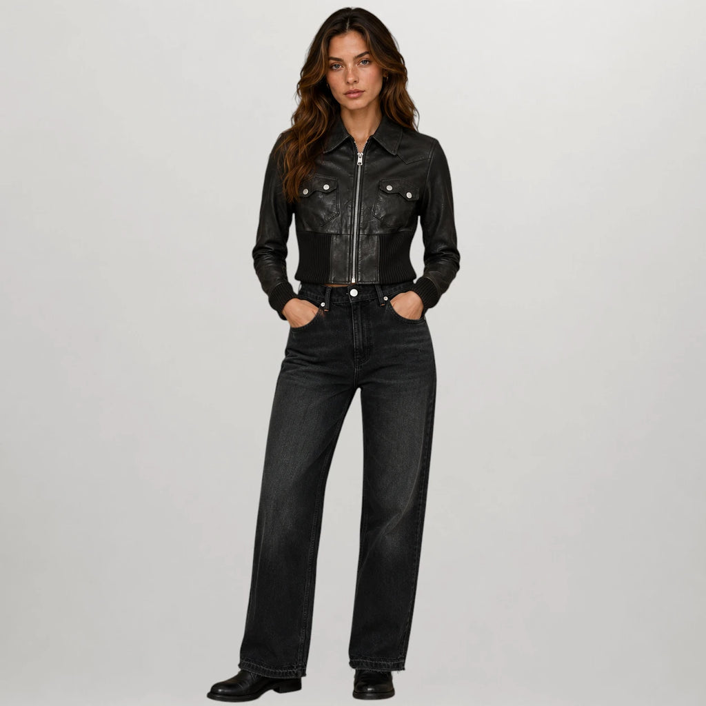 A woman stands against a light gray studio background wearing a black cropped leather jacket with silver hardware and black wide-leg jeans.