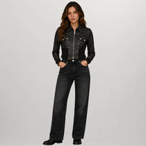 A woman stands against a light gray studio background wearing a black cropped leather jacket with silver hardware and black wide-leg jeans.