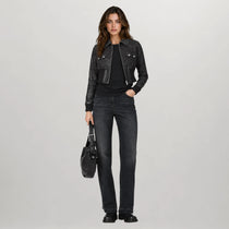 A woman stands against a light gray studio background wearing a black leather moto jacket over a black top and dark gray jeans, holding a black handbag.