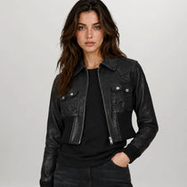 A woman modeling a black leather cropped jacket with silver snap and zipper details, worn over a black top and dark jeans against a light gray studio background.