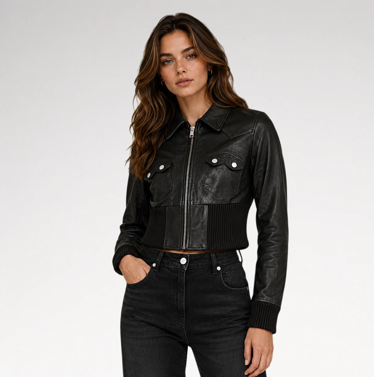 A woman modeling a black leather cropped jacket with a front zipper, ribbed knit trim, and silver snap-button chest pockets, paired with dark jeans against a light gray studio background. Main image
