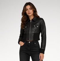 A woman modeling a black leather cropped jacket with a front zipper, ribbed knit trim, and silver snap-button chest pockets, paired with dark jeans against a light gray studio background.