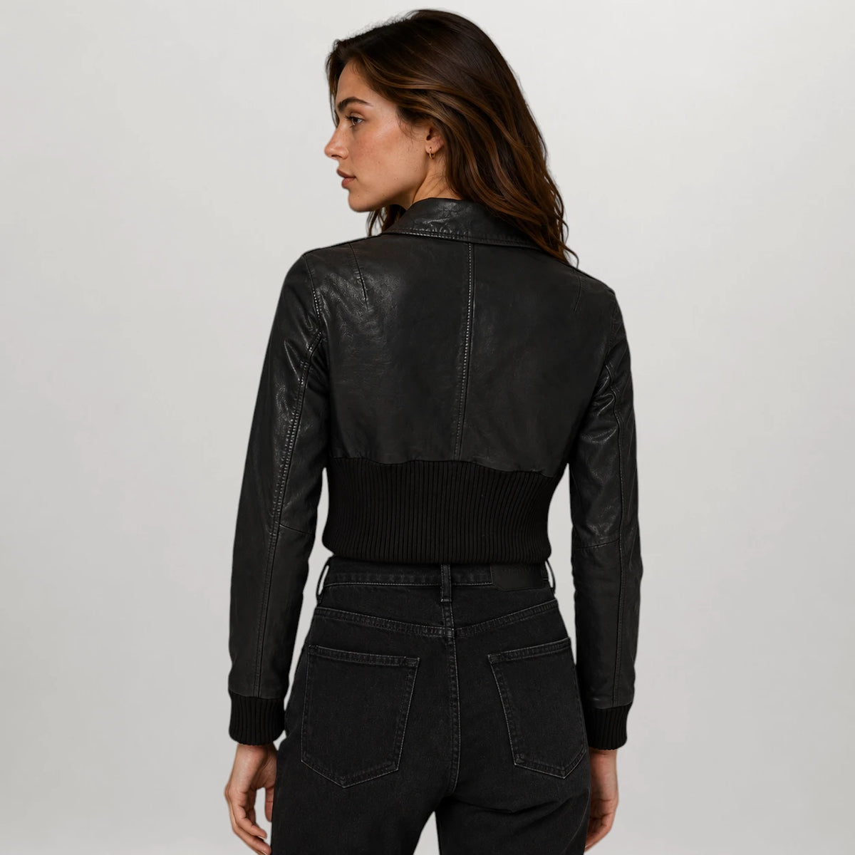 A woman shown from the back wearing a black faux-leather cropped jacket with a ribbed knit hem and cuffs, paired with black jeans, against a light gray studio background.