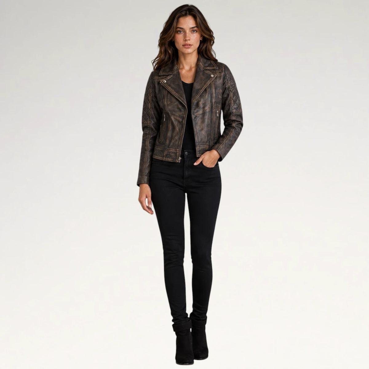 A woman modeling a distressed brown faux-leather moto jacket with gold-tone hardware, paired with black skinny jeans and black ankle boots, against a plain white studio background.