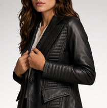 A close-up of a woman wearing a black faux-leather blazer jacket with quilted details on the chest and sleeves, styled over a white top against a light gray background.