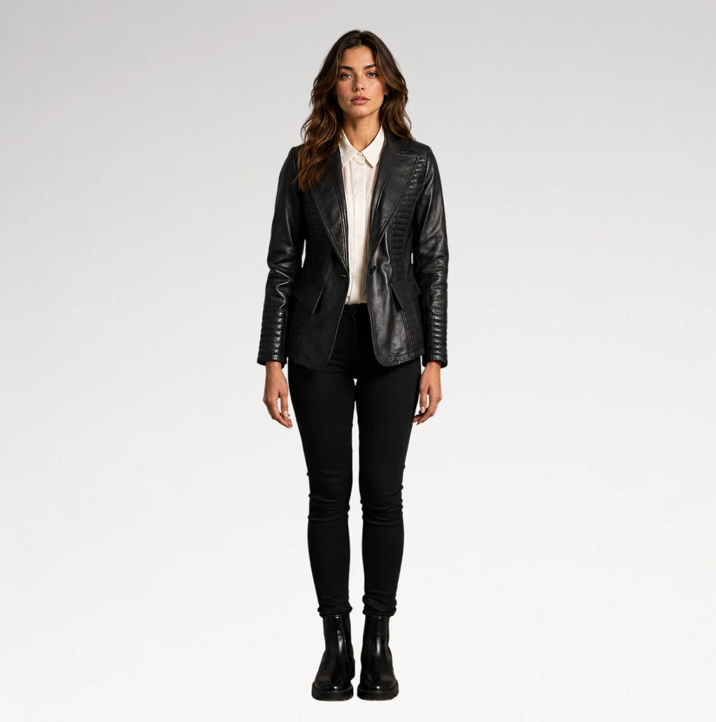 A woman stands against a light gray studio background wearing a black faux-leather blazer with quilted side panels, a white collared shirt, black skinny pants, and black ankle boots.
