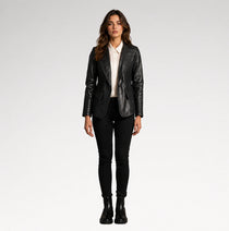 A woman stands against a light gray studio background wearing a black faux-leather blazer with quilted side panels, a white collared shirt, black skinny pants, and black ankle boots.