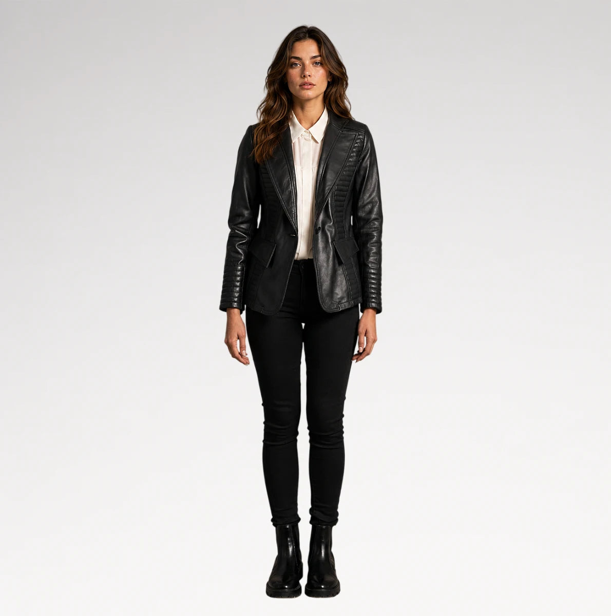 A woman stands against a light gray studio background wearing a black faux-leather blazer with quilted side panels, a white collared shirt, black skinny pants, and black ankle boots.