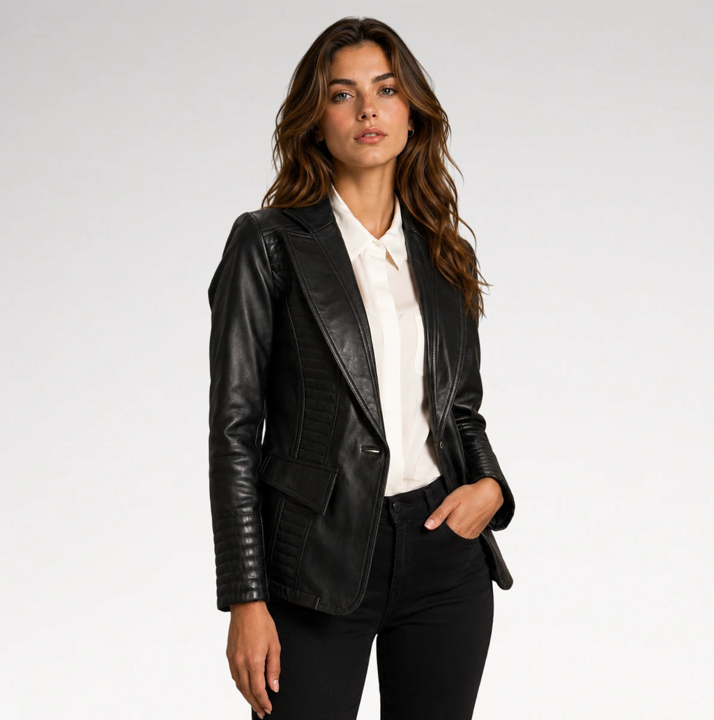 A woman modeling a black faux-leather blazer with quilted panel details, worn open over a white collared shirt and black pants, against a light gray studio background.