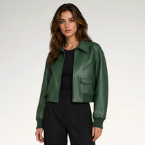 A woman modeling a dark green faux-leather cropped jacket over a black top and black trousers, photographed against a clean light gray studio background.