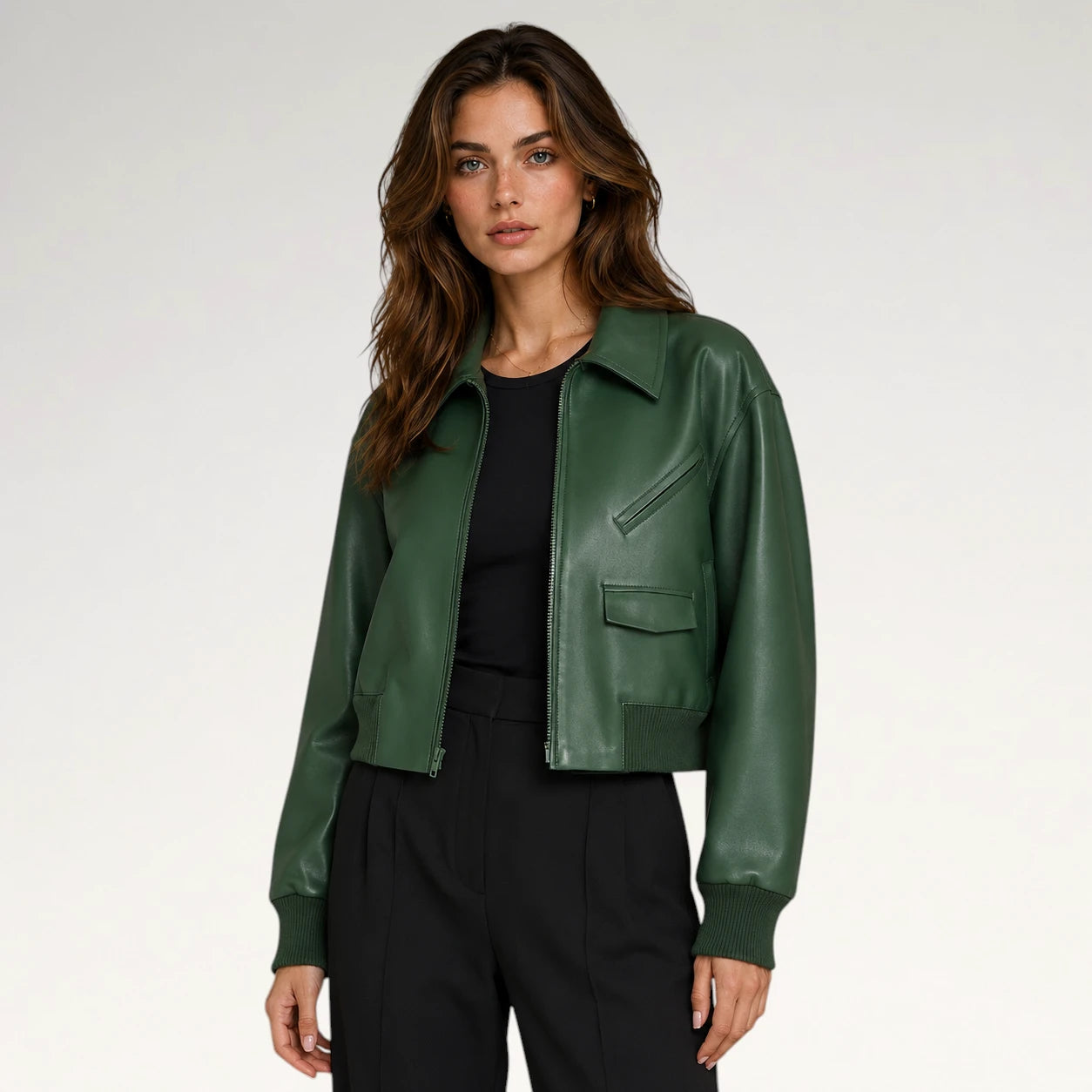 A woman modeling a dark green faux-leather cropped jacket over a black top and black trousers, photographed against a clean light gray studio background.