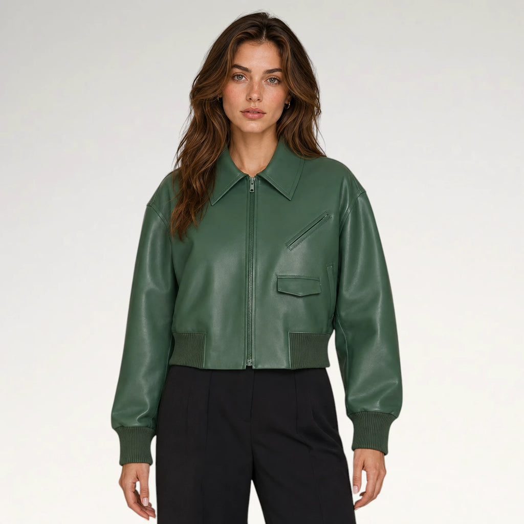 A woman modeling a cropped green faux-leather bomber jacket with a front zipper, ribbed collar, cuffs, and hem, paired with black trousers, against a light gray studio background.