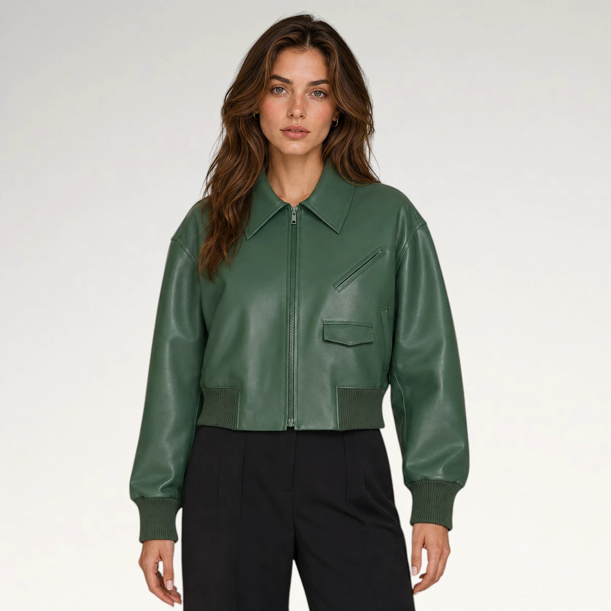 A woman modeling a cropped green faux-leather bomber jacket with a front zipper, ribbed collar, cuffs, and hem, paired with black trousers, against a light gray studio background.