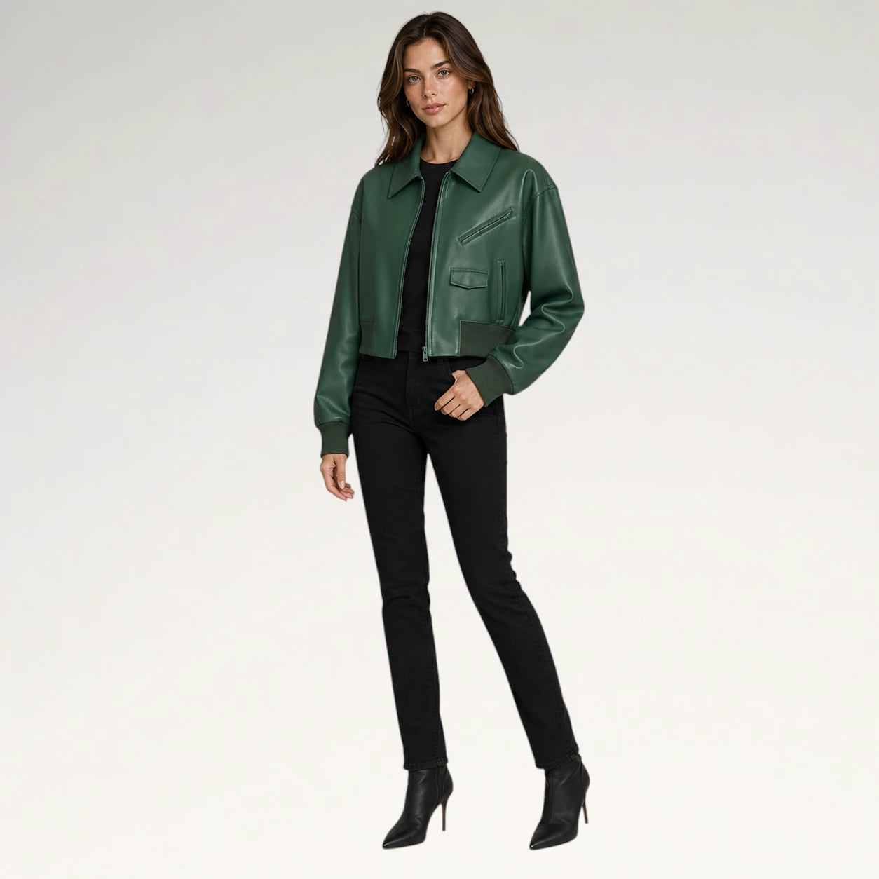 A woman modeling a dark green leather jacket over a black top and black skinny pants, paired with black ankle boots, against a plain light gray studio background.
