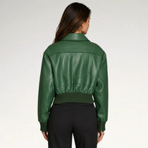 A woman viewed from the back wearing a cropped green faux-leather jacket with a ribbed hem and cuffs, paired with black pants, against a light gray studio background.