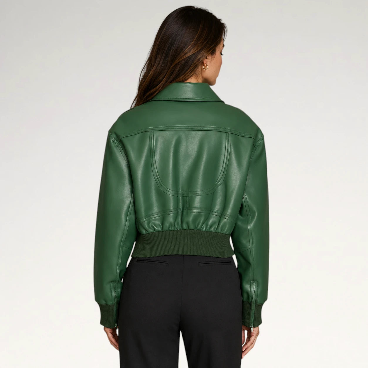A woman viewed from the back wearing a cropped green faux-leather jacket with a ribbed hem and cuffs, paired with black pants, against a light gray studio background.