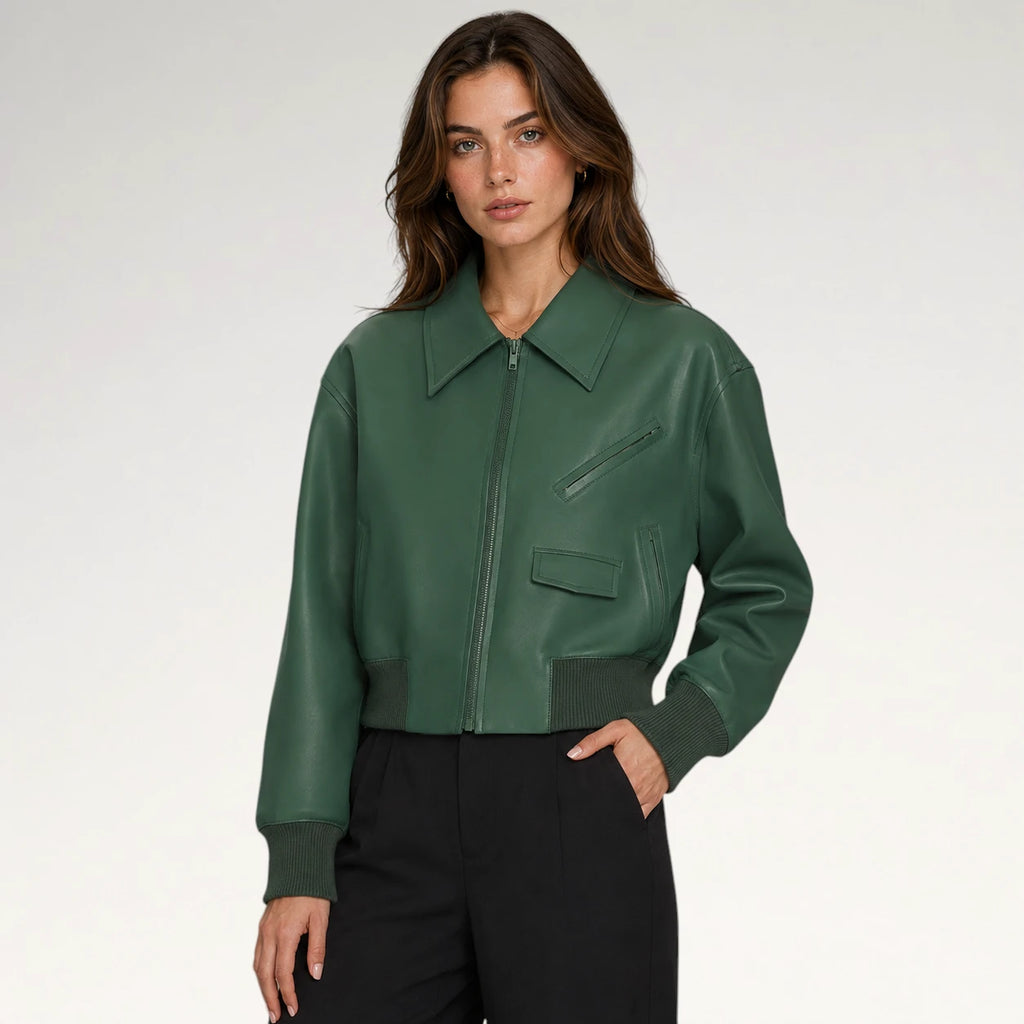 A woman modeling a dark green faux-leather bomber jacket with a pointed collar, zippered front, and diagonal chest pocket, paired with black trousers against a light gray studio background.