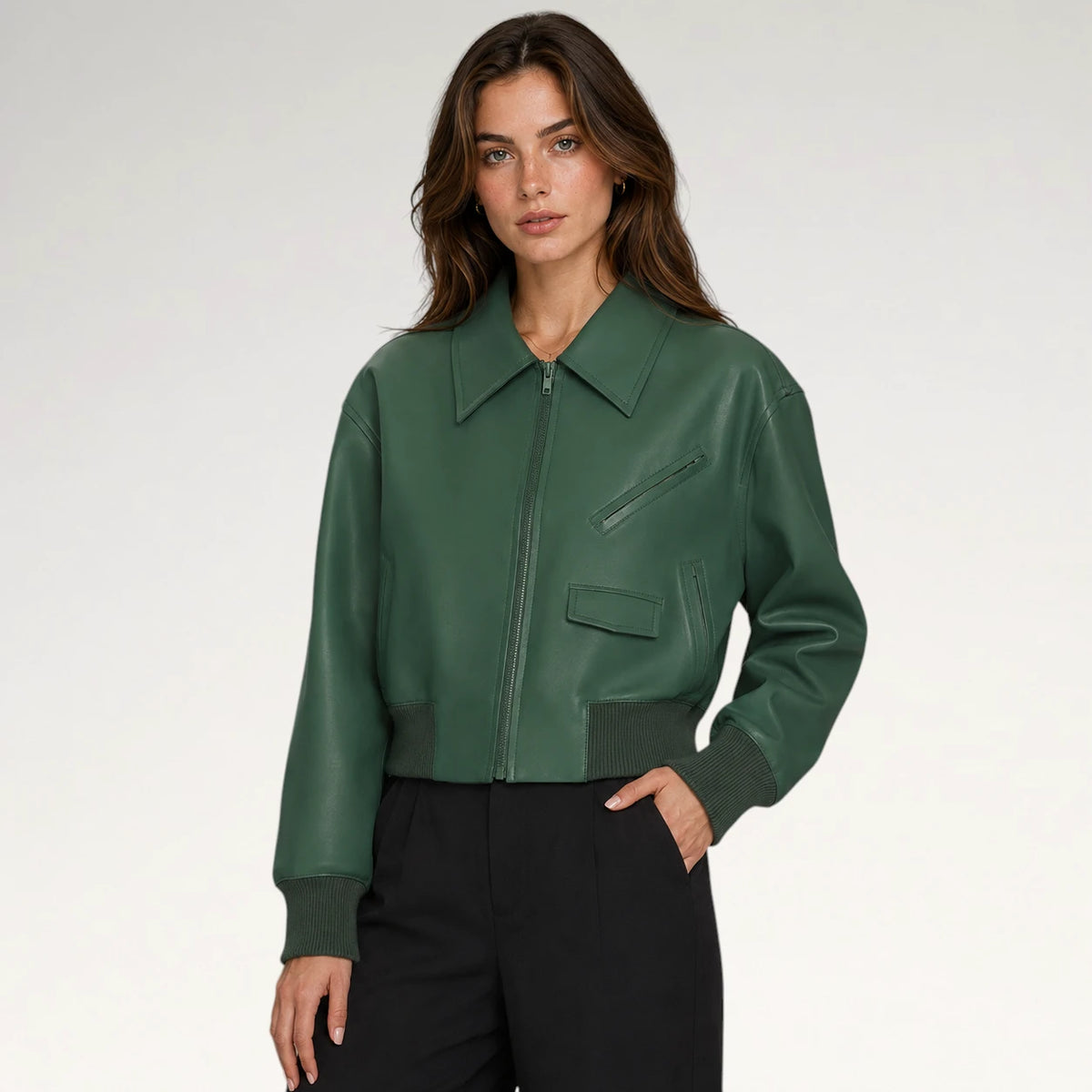 A woman modeling a dark green faux-leather bomber jacket with a pointed collar, zippered front, and diagonal chest pocket, paired with black trousers against a light gray studio background. Main image