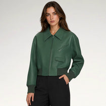 A woman modeling a dark green faux-leather bomber jacket with a pointed collar, zippered front, and diagonal chest pocket, paired with black trousers against a light gray studio background.