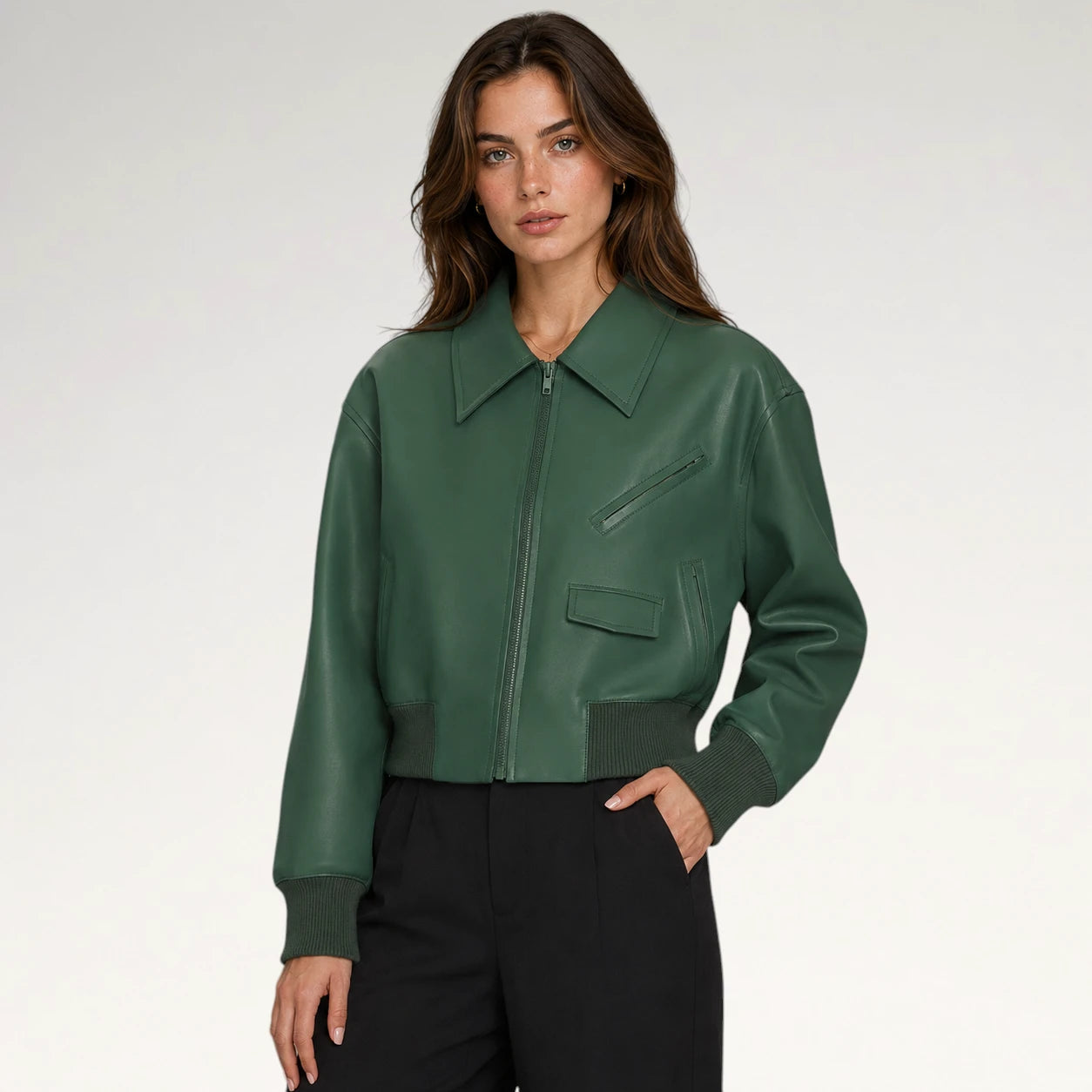 A woman modeling a dark green faux-leather bomber jacket with a pointed collar, zippered front, and diagonal chest pocket, paired with black trousers against a light gray studio background.