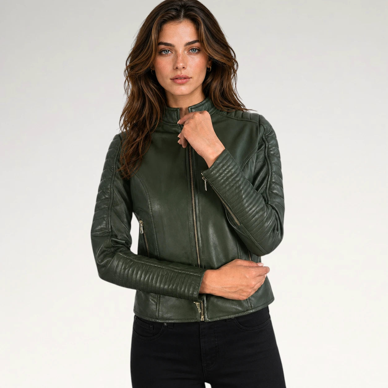 A woman modeling a dark olive green faux leather biker jacket with gold-tone zippers, paired with black jeans, against a plain light gray studio background.