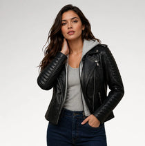 A woman modeling a black faux-leather moto jacket with a gray hood, worn over a gray tank top and dark blue jeans, against a light gray studio background.