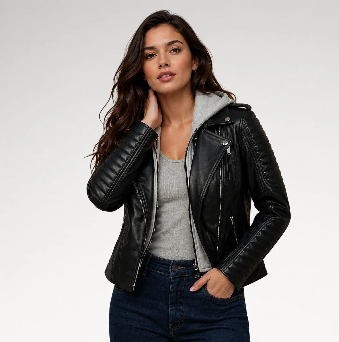 A woman modeling a black faux-leather moto jacket with a gray hood, worn over a gray tank top and dark blue jeans, against a light gray studio background.
